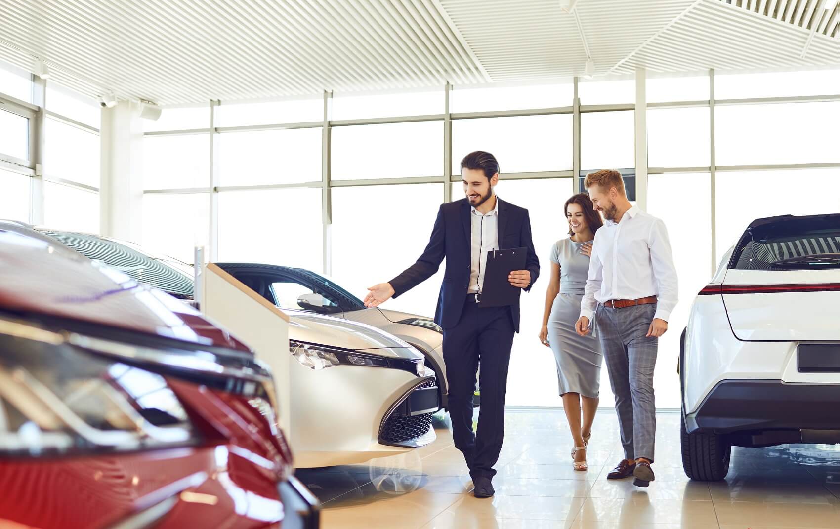 A bright, modern used car dealership showroom features a salesperson presenting vehicles to a couple. Several new cars are displayed throughout the spacious showroom, creating a welcoming setting for exploring different vehicle options.